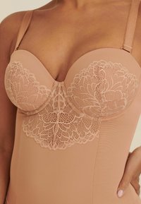 Nude bodysuit met een kantoverlay op de buste, een gladde textuur en verstelbare bandjes. De gebogen vorm accentueert de figuur en biedt ondersteuning.