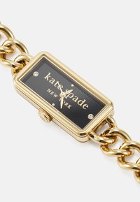 Złoty prostokątny zegarek z czarną tarczą, z nazwą marki "kate spade" i dwoma małymi kryształkami, przymocowany do masywnej bransoletki łańcuszkowej.