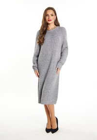 Abito maglione grigio a maglia con texture a coste, maniche lunghe e tasche laterali, che arriva a metà polpaccio, abbinato a tacchi alti neri.