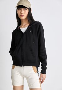 Un sweat à capuche noir zippé avec des cordons de serrage, présentant un petit logo, porté par-dessus un haut blanc et associé à un short ajusté beige et noir.
