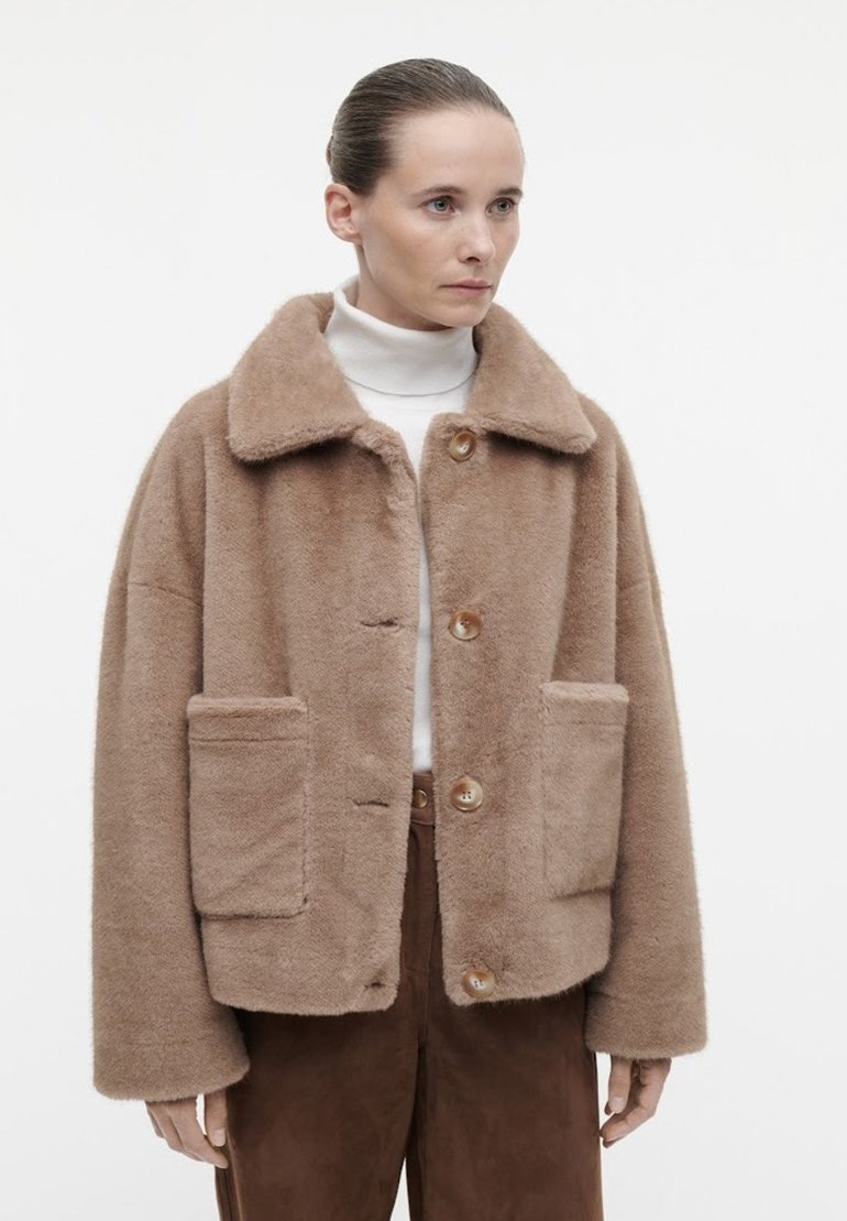 Braune, flauschige Außenjacke mit Kragen, zwei Vorderaschen und großen Knöpfen, kombiniert mit einem weißen Rollkragenpullover darunter.