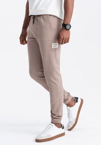 Beżowe spodnie dresowe o fakturowanym wykończeniu, z dwoma bocznymi kieszeniami, elastycznym paskiem z trokiem i metką na lewej udzie. W zestawie z białymi sneakersami.