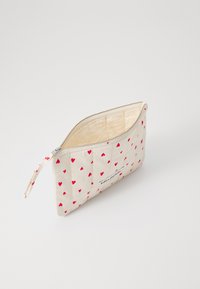 Quilted cosmetic pouch in cream fabric with small red hearts. Zipper closure with a wrist strap. Interior is beige. Features a logo label.