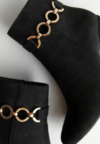 Botines de ante negro con acentos de cadena de metal dorado. Presentan un diseño elegante, punta afilada y un tacón alto. Acabado mate texturizado.