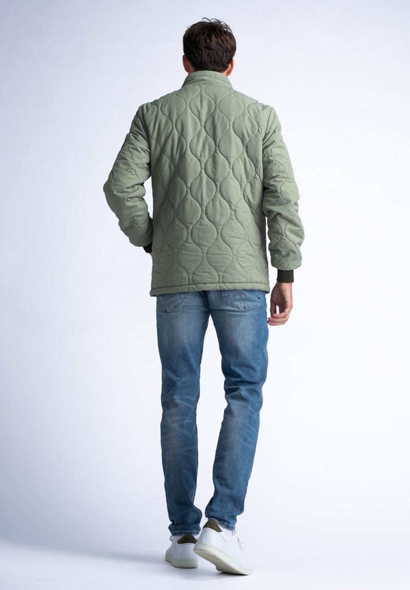 Chaqueta acolchada de color verde oliva con un alto cuello y puños acanalados, llevada sobre jeans de color azul claro y zapatillas blancas, vista desde atrás.