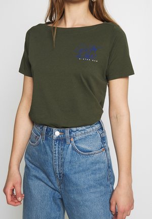 Kobieta w oliwkowej koszulce z krótkim rękawem z niebieskim logo "G-Star Raw" w połączeniu z jasnoniebieskimi jeansami z wysokim stanem.