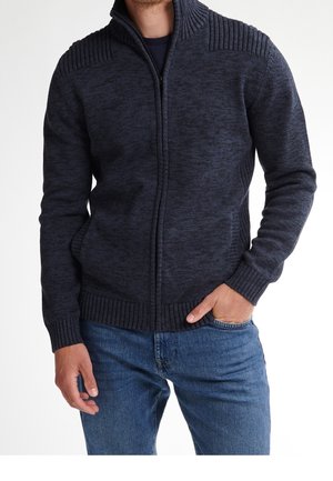 Mann trägt einen dunkelmarineblauen Strickpullover mit Reißverschluss, geripptem Kragen, Bündchen und Saum, kombiniert mit blauen Jeans, eine Hand in der Tasche.