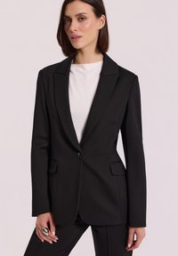 Kobieta z krótkimi brązowymi włosami, ubrana w czarną, skrojoną marynarkę na białą bluzkę, czarne spodnie i duży srebrny pierścionek na lewej ręce.