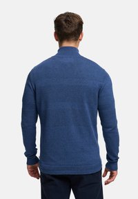 Blå stickad tröja med hög krage, strukturerat mönster med horisontella ränder och ribbade muddar. Materialet verkar mjukt och slitstarkt.