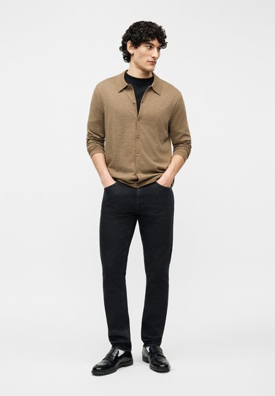 Νέος άνδρας με σγουρά μαλλιά, φορώντας ένα καφέ ζακέτα με κουμπιά πάνω από μαύρο πουκάμισο, μαύρο τζιν και μαύρα δερμάτινα παπούτσια, με τα χέρια στις τσέπες, κοιτάζει προς τα δεξιά.