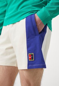 Homme portant un short crème et bleu avec la main dans la poche latérale, avec un petit patch logo Nike multicolore sur la partie bleue.