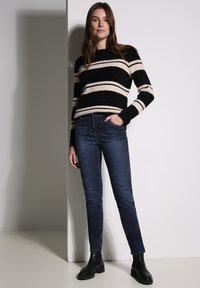 Suéter a rayas negro y crema con cuello redondo, combinado con jeans azul oscuro y botas negras de tobillo. La modelo está de pie frente a un fondo liso.