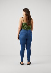 Jeans ajustados azules combinados con una blusa verde estampada, con tirantes finos y un diseño ceñido, visto desde la parte trasera, mostrando dos bolsillos traseros.