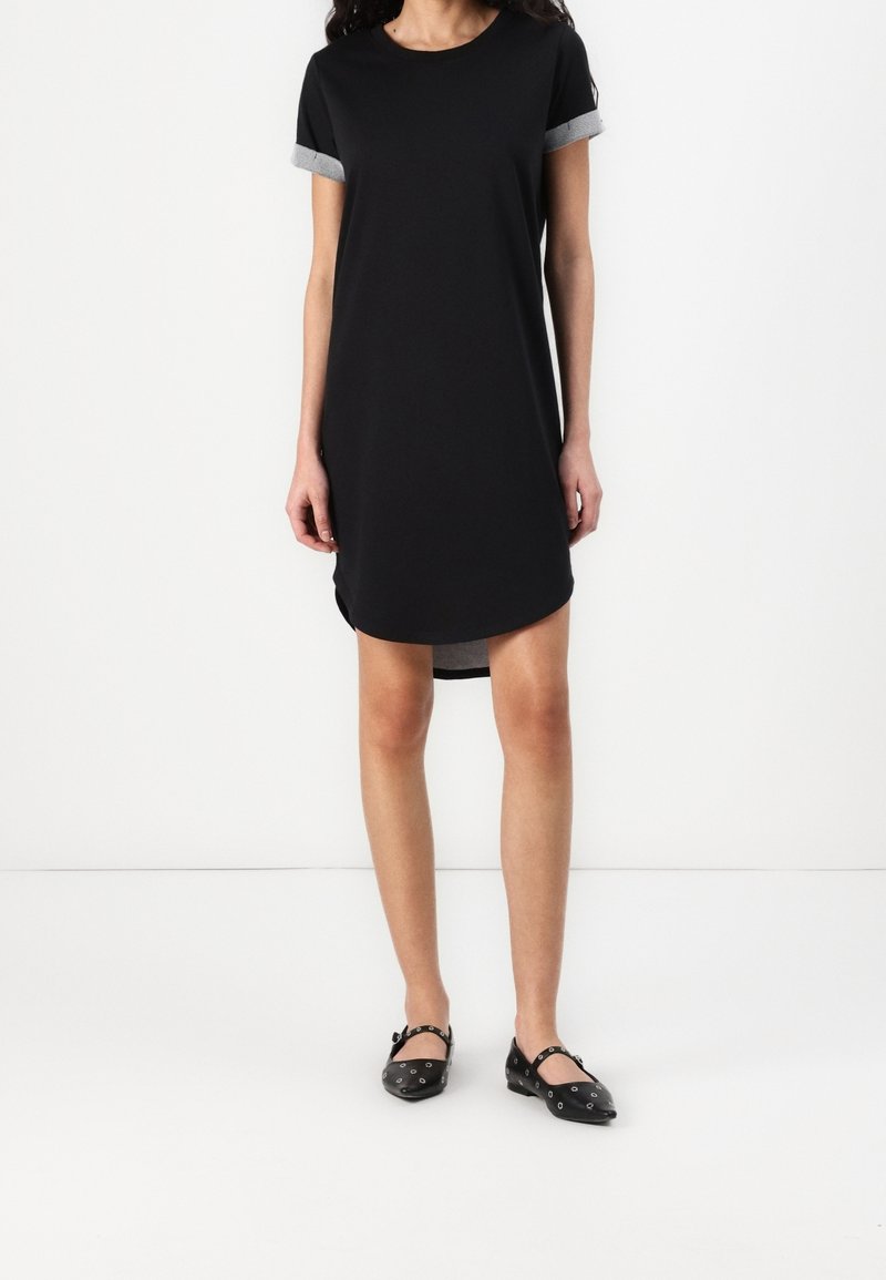 Femme portant une robe noire à manches courtes avec des poignets gris roulés et des chaussures plates noires avec des œillets argentés, debout sur un sol blanc.