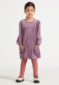 Lila Velourskleid mit langen Ärmeln, Blumenstickerei und zwei Taschen, kombiniert mit rosa gerippten Leggings und cremefarbenen Schuhen.