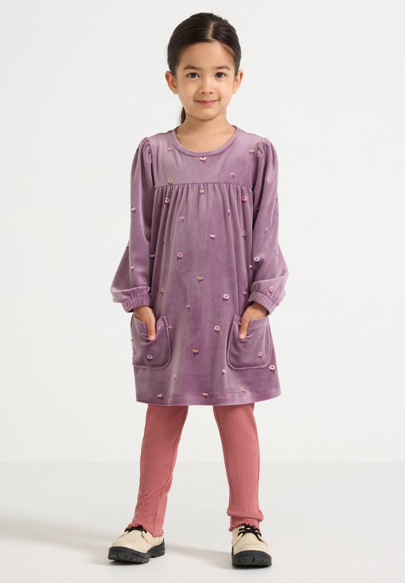 Lila Velourskleid mit langen Ärmeln, Blumenstickerei und zwei Taschen, kombiniert mit rosa gerippten Leggings und cremefarbenen Schuhen.