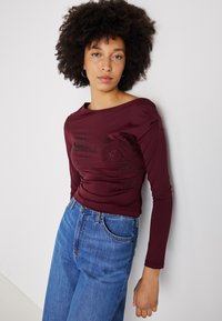 Līks sarkans garām piedurknēm tops ar laivu kaklu, ar dekoratīviem akcentiem priekšpusē, apvienots ar augsto jostasvietu ziliem džinsiem.