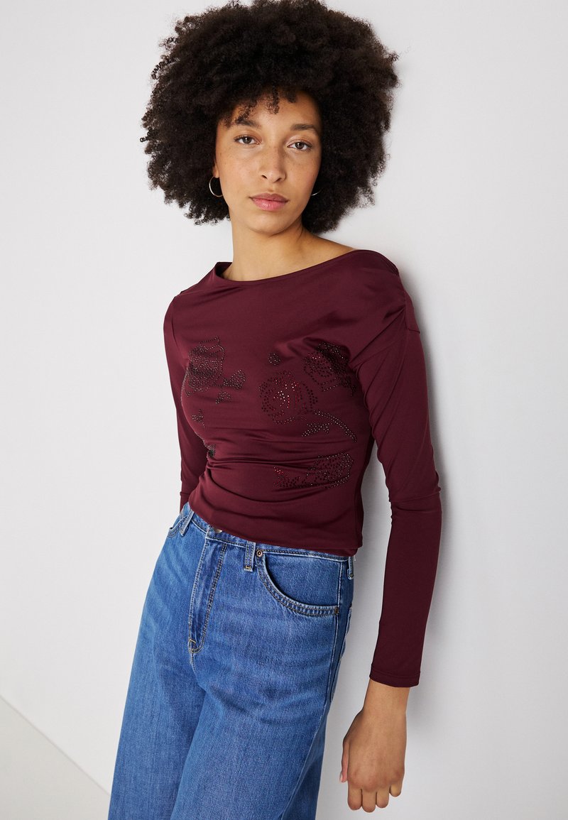 Līks sarkans garām piedurknēm tops ar laivu kaklu, ar dekoratīviem akcentiem priekšpusē, apvienots ar augsto jostasvietu ziliem džinsiem.