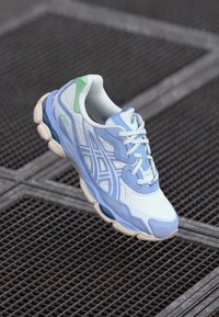 Lyseblå og hvide sneakers med grønne accenter, mesh- og ruskindsstrukturer, fremtrædende snørebånd og en mønstret mellemsål.