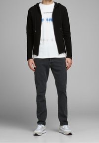 Černá mikina na zip s texturovaným povrchem, přes bílý batikovaný tričko s modrými akcenty, doplněná tmavými džínami a bílými teniskami.