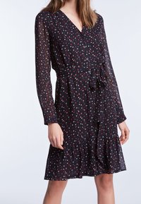 Zwarte jurk met kleine rode en witte hartpatronen, lange doorschijnende mouwen, V-hals, knopen aan de voorkant, tailleband en ruches aan de onderkant.