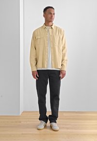 Camisa de pana beige, camiseta blanca debajo, pantalones negros y zapatillas crema. Suelo de madera natural, fondo de pared neutro.