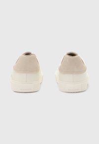 Beige Wildleder- und weiße Leder-Sneaker mit niedrigem Profil, gepolstertem Absatz und strukturiertem Gummisohle. Branding auf der Rückseite sichtbar.