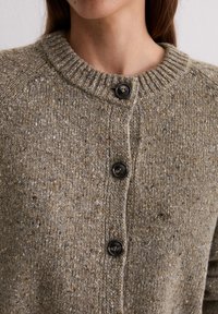 Nahaufnahme einer gefleckten braunen Strickjacke mit einem gerippten Rundhalsausschnitt und drei dunklen Knöpfen, die an einer Person zugeknöpft ist.