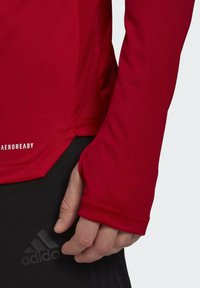 Rotes langärmliges Sportshirt aus leichtem Material, mit enger Passform und Daumenlöchern an den Bündchen. Markenlogo auf der Hose sichtbar.