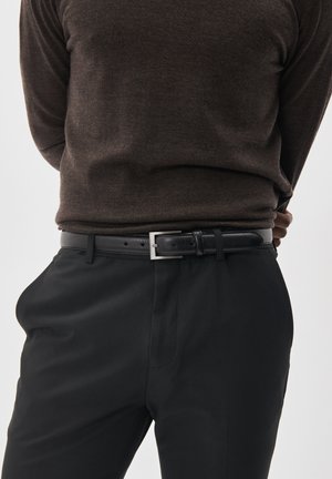 Cinturón de cuero negro con una hebilla cuadrada plateada, llevado sobre pantalones negros. El suéter de punto es marrón oscuro, ajustándose al cuerpo.