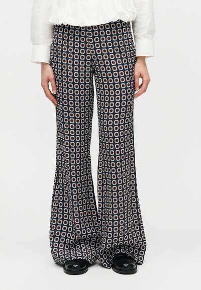 Μαύρο παντελόνι με φαρδιά μπάτζα και γεωμετρικό λευκό και καφέ λουλουδάτο σχέδιο, φορεμένο με μαύρα παπούτσια και λευκό μπλουζάκι με μακριά μανίκια.