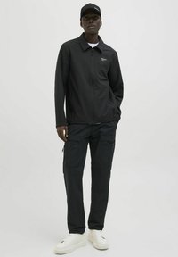 Schwarze Jacke mit Kragen und Reißverschluss, kombiniert mit schwarzen Cargo-Hosen mit mehreren Taschen und weißen Plateau-Sneakern.