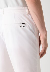 Pantalons blancs au toucher lisse avec une poche zippée à l'arrière. Présente un logo "BOSS" noir sur la poche.