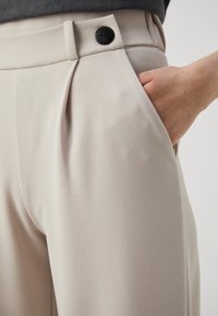 Pantalons plissés beiges en tissu texturé, avec une fermeture à bouton noir et une poche latérale, montrés de la taille à la hanche.