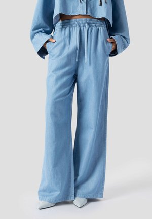 Femme portant un pantalon en denim bleu clair à jambes larges avec une taille élastique et un cordon de serrage, assorti à des chaussures blanches à bout pointu.