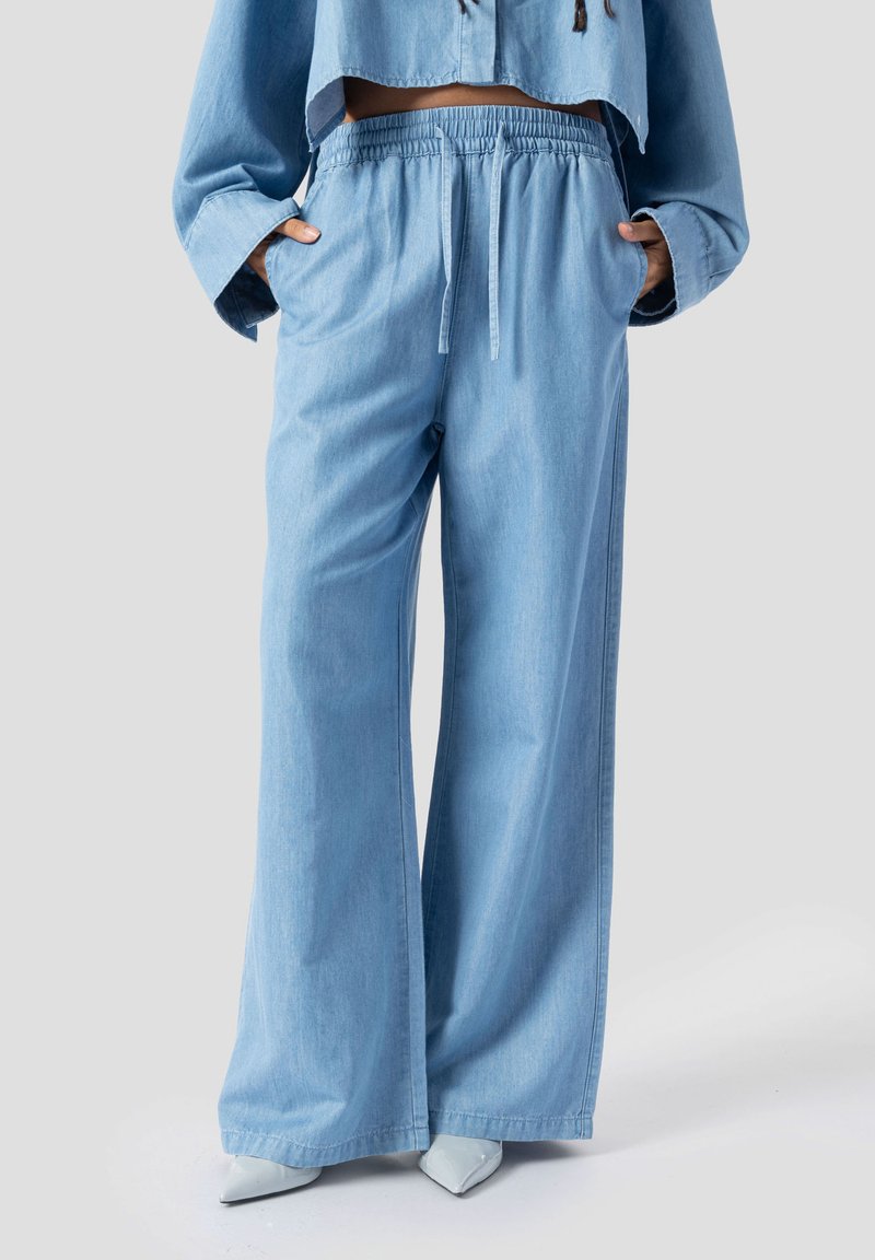 Femeie purtând pantaloni de blugi largi, de culoare albastru deschis, cu talie elastică și șnur, asortați cu pantofi albi cu vârf ascuțit.