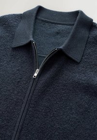 Tmavomodrá fleecová bunda na zips s textúrovaným povrchom, ribovaným golierom a detailom kovového zipsu. Jednoduchý design s teplým vzhľadom.