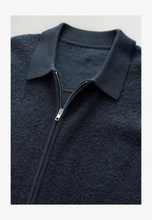 Tmavě modrá fleecová bunda na zip s texturovaným povrchem, žebrovaným límcem a kovovým detailem zipu. Jednoduchý design s teplým vzhledem.