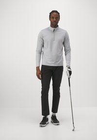 Lys grå langermet topp med kvart glidelås og ribbestruktur, kombinert med svarte golfbukser. Modellen holder en golfkølle, iført svarte sko og hansker.