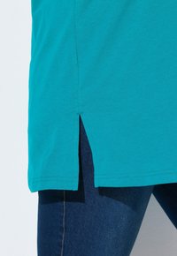 Tunica in cotone turchese con spacco laterale e orlo dritto, abbinata a jeans blu scuro. Le cuciture a vista migliorano il dettaglio strutturale.