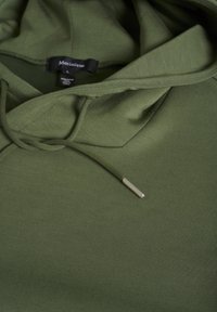 Sudadera de color verde oliva con capucha ajustable, textura suave y una pequeña punta metálica en el cordón; etiqueta de la marca visible en el interior.