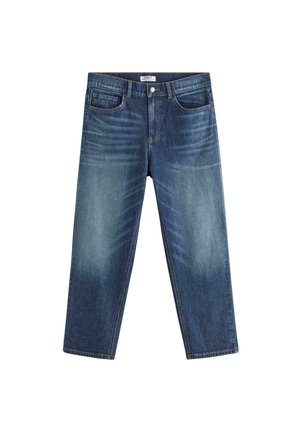 Smilende dreng med kort blondt hår står mod en ensfarvet baggrund iført en hvid t-shirt, løse blå jeans og hvide sneakers.