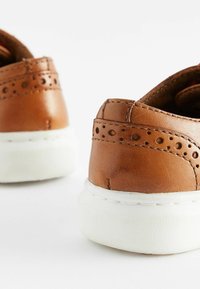 Baskets en cuir marron avec des accents perforés, semelles en caoutchouc blanc et détails de couture. La vue arrière montre des talons ronds et une texture lisse.