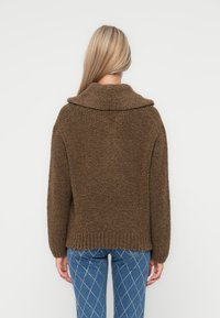 Brun strikket genser med en bred krage, strukturert stoff og ribbestrikket kant. Kombinert med blå bukser med diamantmønster. Vist fra bakdelen.