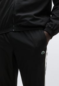 Mustat urheiluhousut, jotka on valmistettu kevyestä kankaasta. Housuissa on vihreä logoyksityiskohta ja sivuilla kontrastiväriset raidat lisäyksityiskohtina.