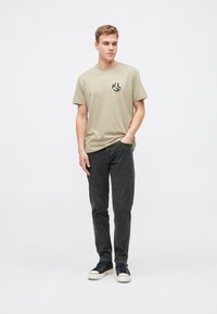 Camiseta verde oliva claro con un logo negro en el pecho, combinada con jeans gris oscuro y zapatillas negras con suelas blancas.