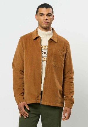 Homme portant une veste en velours côtelé marron par-dessus un pull à col roulé blanc à motifs et un pantalon vert, debout devant un fond clair uni.