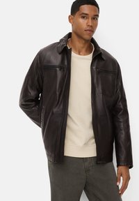 Donkerbruine leren jas met ritszakken, een staande kraag en een licht gestructureerd oppervlak. Draagt over een lichtbeige top.