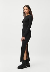 Vestido preto de mangas longas com rachas laterais, silhueta ajustada, feito de um tecido suave. Usado com botas pretas de solado grosso.
