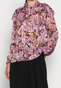 Top trasparente con motivo floreale in tonalità di rosa, viola e marrone. Presenta maniche arricciate e una scollatura a chiave. Taglio leggermente ampio.